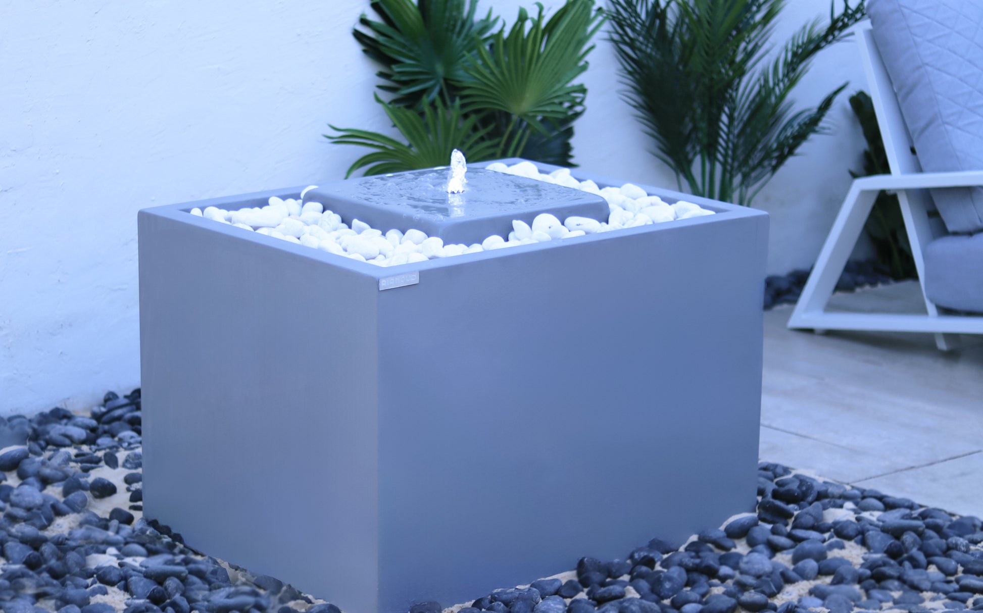 نافورة حديثة مستطيلة الشكل مصنوعة من الخرسانة بلمسة نهائية رمادية، محاطة بالحصى عند القاعدة، مزوّدة بإضاءة داخلية، ومموضعة في الفناء بجانب كرسي استرخاء.