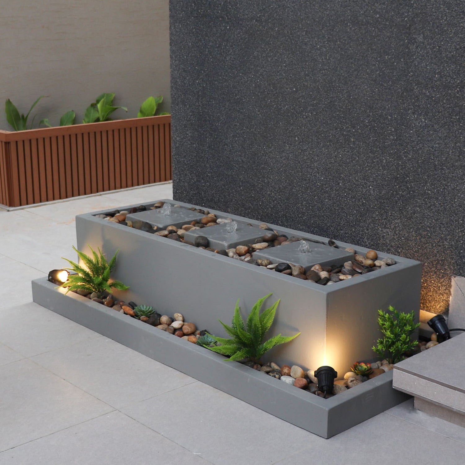 نافورة داخلية عصرية، بقاعدة خرسانية مستطيلة باللون الرمادي مزوّدة بإضاءة مدمجة وأحجار زخرفية، محاطة بمساحة ذات جدران رمادية وفاصل خشبي في الخلفية.