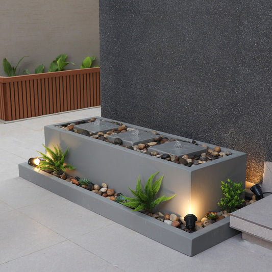 نافورة داخلية عصرية، بقاعدة خرسانية مستطيلة باللون الرمادي مزوّدة بإضاءة مدمجة وأحجار زخرفية، محاطة بمساحة ذات جدران رمادية وفاصل خشبي في الخلفية.