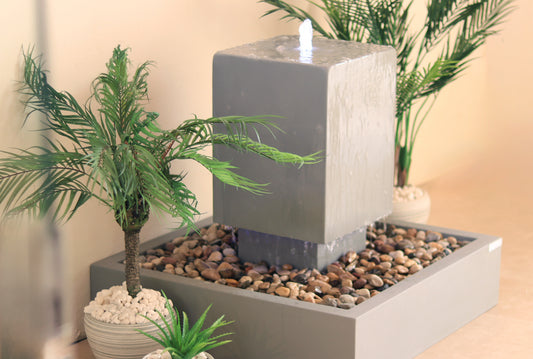 نافورة داخلية بتصميم عصري، تحتوي على حوض سفلي مربع مملوء بالصخور، تعلوه كتلة خرسانية مكعبة. تحيط بالنافورة نباتات مزروعة في أصص تضيف لمسة من الخضرة.