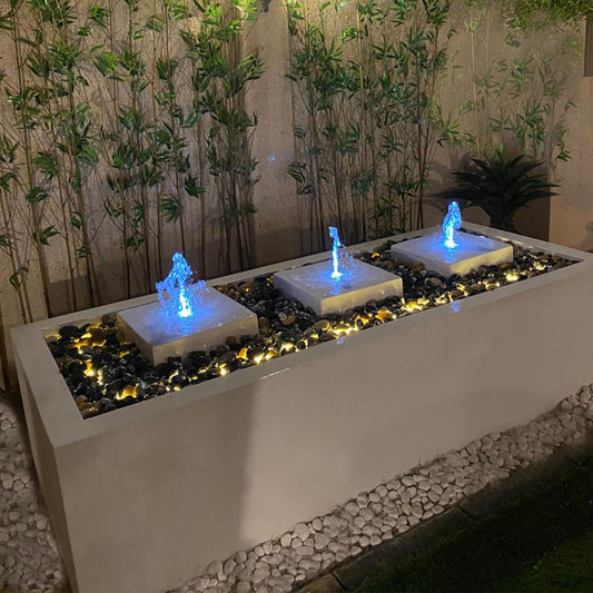 نافورة خارجية تحتوي على ثلاث شلالات مياه مضاءة باللون الأزرق، موضوعة داخل حوض مستطيل من الخرسانة ومحاطة بأحجار زخرفية، مع وجود نباتات خضراء في الخلفية